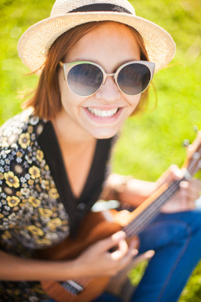 Attractive young woman wearing sunglasses and straw hat playing on brown wooden Ukulele outdoors. Talented students. Music lessons. Traditional musical instruments. Ethnic music. Music festival, open air.