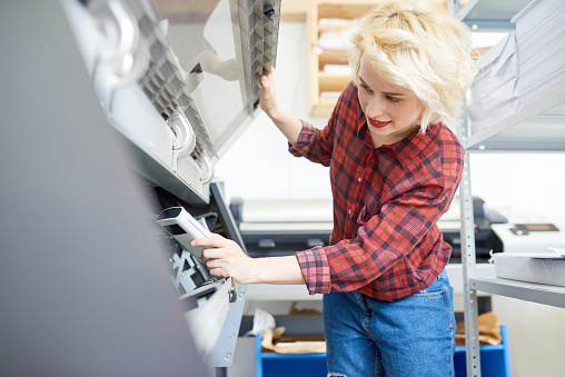 Beautiful young woman in casual outfit inspecting plotter while working in printing office.