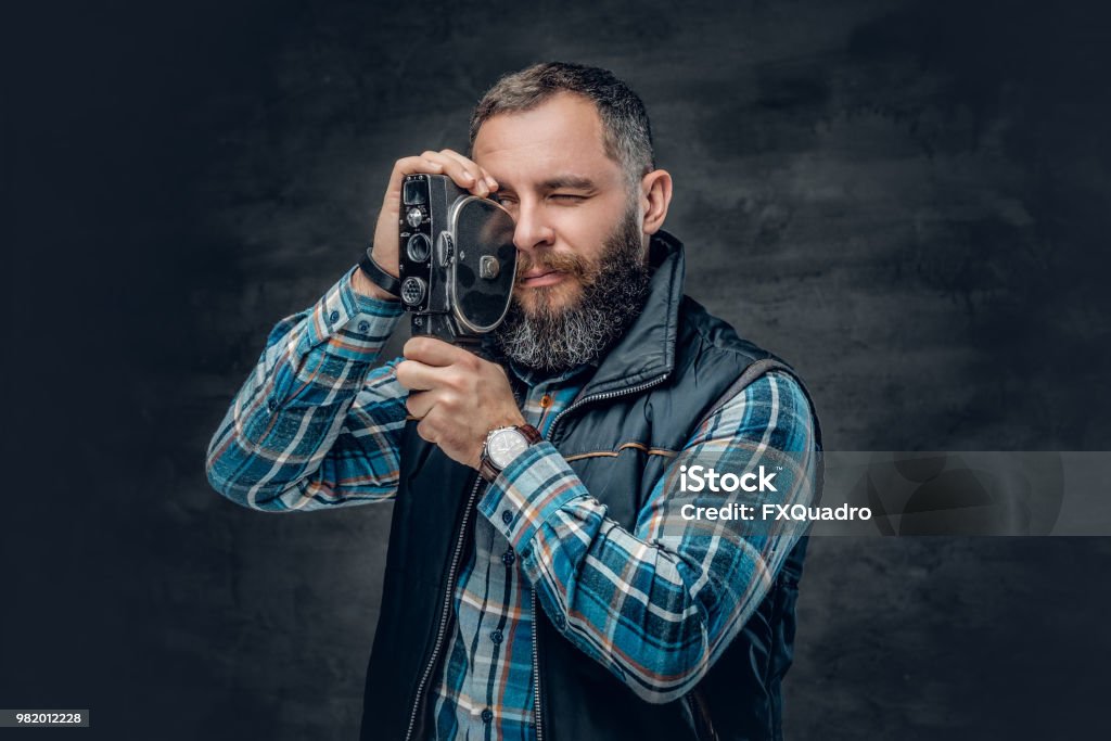 Bearded middle age male holds vintage 8 mm video camera. Portrait of bearded middle age male holds vintage 8 mm video camera over grey background. Camera Operator Stock Photo