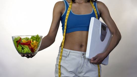 Woman in loose pants standing with scales, holding out bowl of salad, dieting