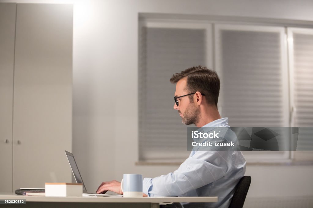 Mature man working on laptop in office Side view of mature man sitting at his desk and working on laptop. Businessman using laptop in office. Adult Stock Photo