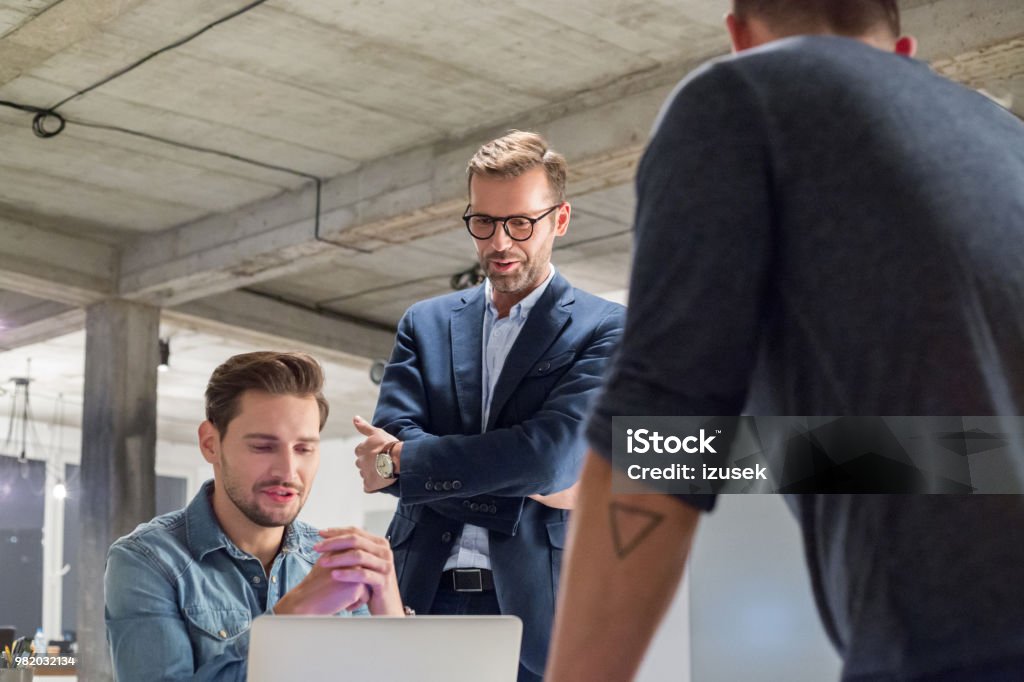 Team of professional discussing work in office Team of professional discussing work in office. Young man sitting at his desk with colleagues standing by. Adult Stock Photo