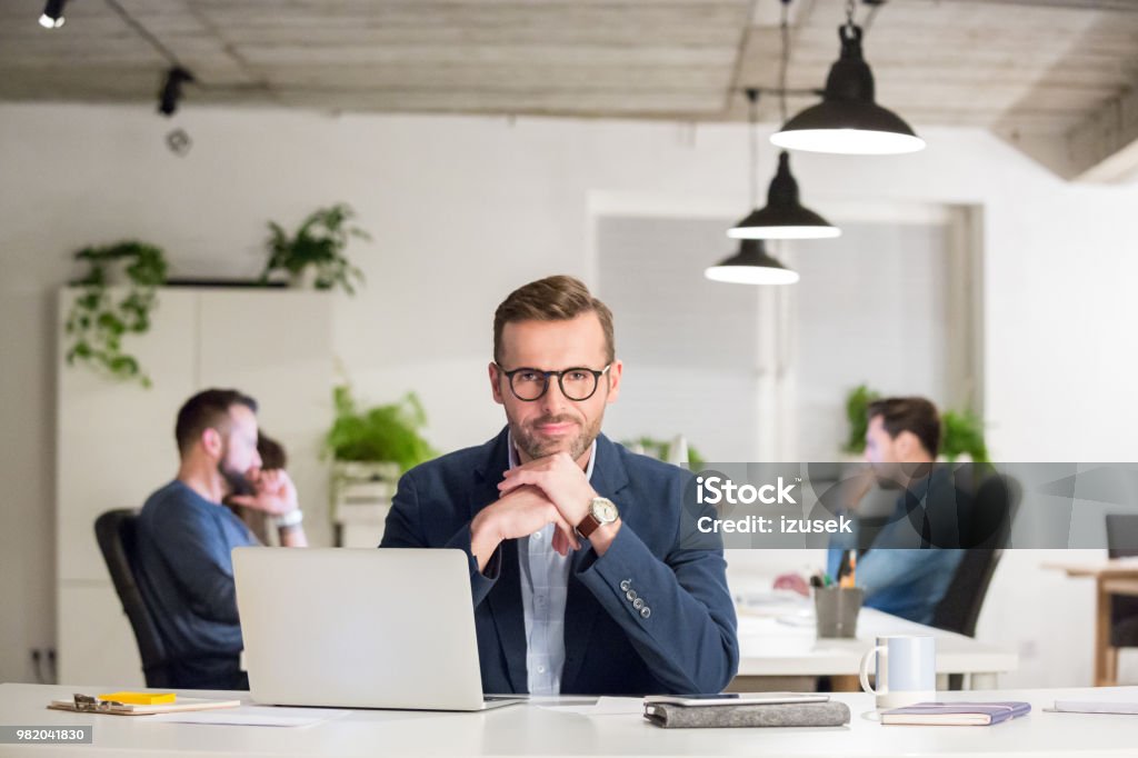Mature businessman sitting at his desk Portrait of confident mature businessman sitting at his desk with his team working in background. Office Stock Photo