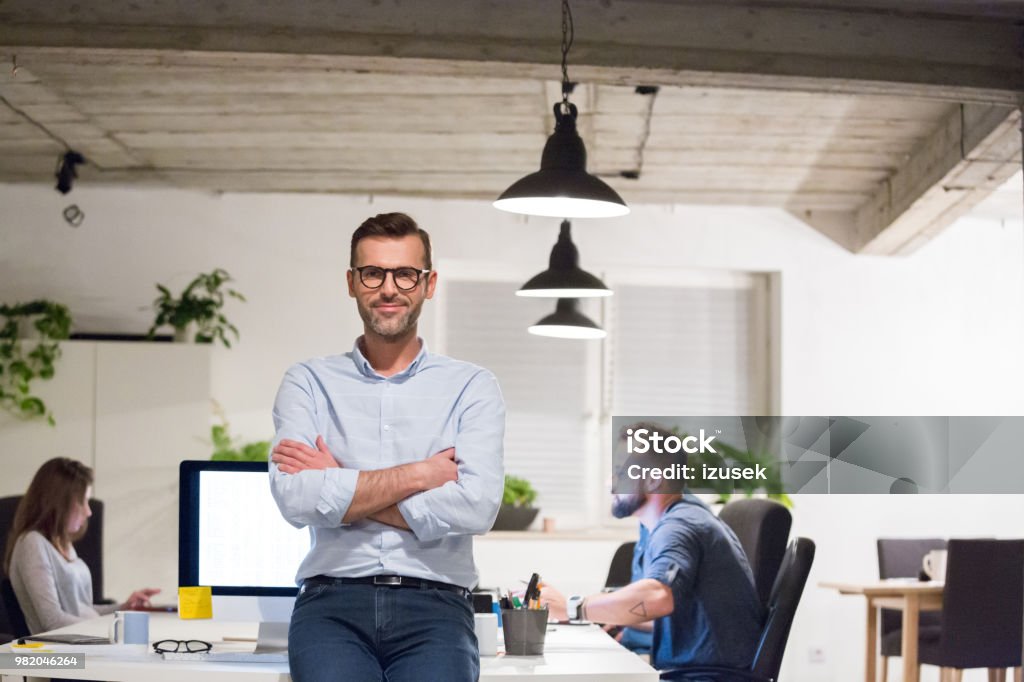 Entrepreneur at his startup office Portrait of handsome mature businessman standing by a desk with coworkers working in background. Entrepreneur standing at his startup office. Adult Stock Photo