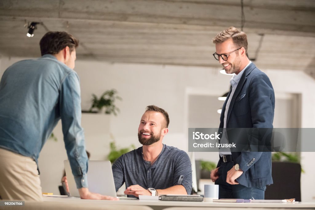 Team of executives working in office Team of executives in office working together. Man sitting at tablet  with laptop and colleagues standing by. Adult Stock Photo
