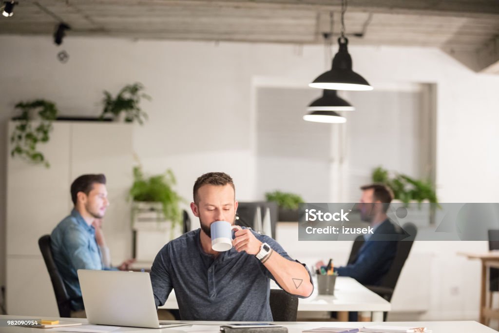 Man drinking coffee at his office desk Mature businessman sitting at his desk and drinking coffee with colleagues working in background. Computer Stock Photo