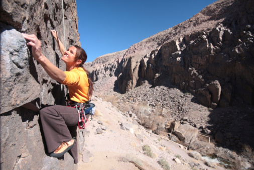 Back view of young woman climbing on cliff rock with blue sky in the background.