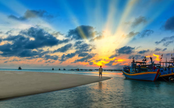 Mui Ne, Vietnam - April 22, 2018: The lonely man watches the sunset on a sandy beach near the fishing village with many boats anchored in distant waters near Mui Ne, Vietnam