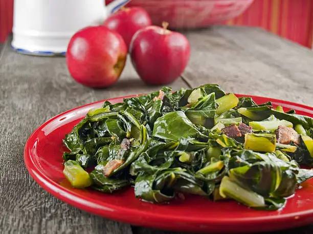 Collard greens and bacon on a red plate with apples and a water pitcher in the background.
