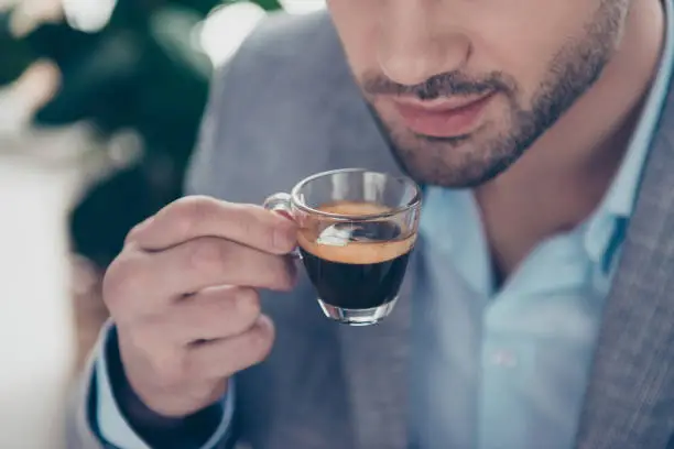 Cropped close up half face portrait of stylish attractive man holding small glass with espresso near mouth, every morning ritual before work in work place