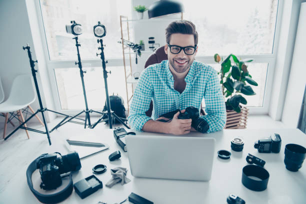 portrait of cheerful positive man in shirt with modern hairstyle, sitting at desk, holding digital camera, looking at camera. electronic devices computer laptop equipments concept - office tool flash imagens e fotografias de stock