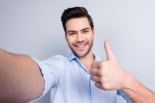 Self portrait of stylish cheerful guy making self picture on front camera and showing thumb up symbol, isolated on grey background