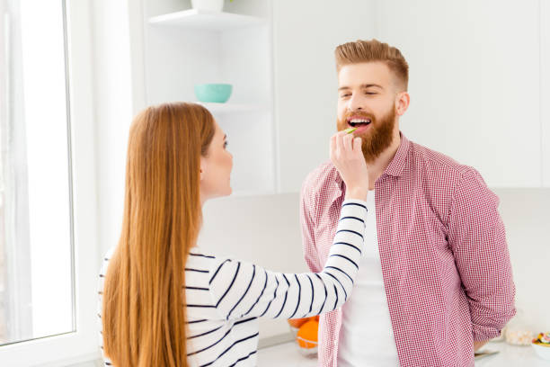 domestic domicile indoors weekend vacation holiday concept. rear view portrait of girl with long red hair giving slice of cucumber to handsome lover keeping mouth open - long hair red hair women men imagens e fotografias de stock