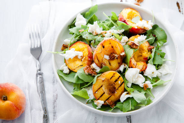 insalata fresca con metà pesca alla griglia, rucola e burrata su un piatto su sfondo bianco in legno in difficoltà. vista dall'alto. concetto di cibo estivo - plate salad leaf vinegar foto e immagini stock