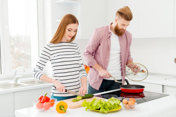 istock Breakfast supper dinner kitchen ware concept. Portrait of busy concentrated partners cooking meal together, beautiful woman cutting cucumber handsome man frying food in modern white kitchen 982423360
