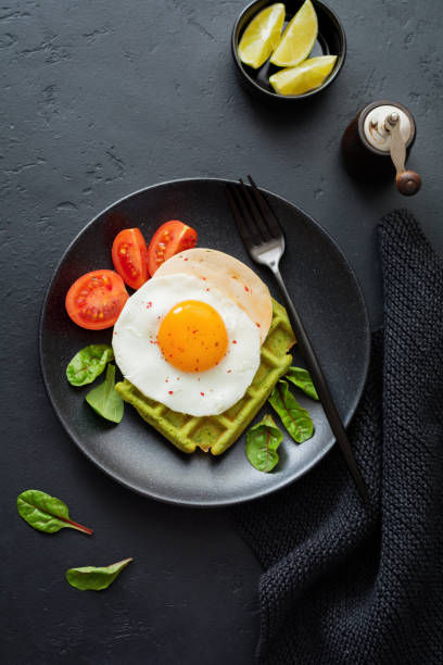 istock Fried egg with cheese, tomatoes, chard, and waffle with spinach on black ceramic plate on dark concrete background. Sandwich for breakfast. Selective focus. Top view. Copy space. 982486646