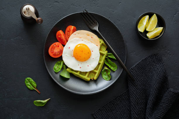 istock Fried egg with cheese, tomatoes, chard, and waffle with spinach on black ceramic plate on dark concrete background. Sandwich for breakfast. Selective focus. Top view. Copy space. 982486672