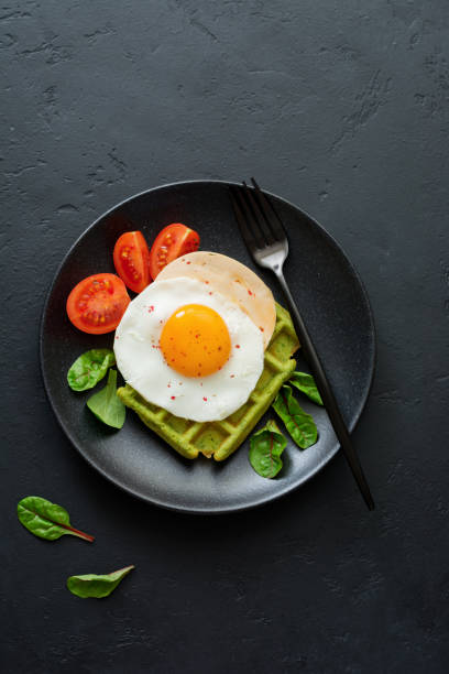 istock Fried egg with cheese, tomatoes, chard, and waffle with spinach on black ceramic plate on dark concrete background. Sandwich for breakfast. Selective focus. Top view. Copy space. 982487384