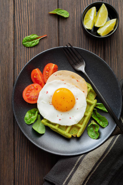 istock Fried egg with cheese, tomatoes, chard, and waffle with spinach on black ceramic plate on brown old wooden background. Sandwich for breakfast. Selective focus. Top view. Copy space. 982487858