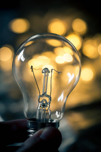 Close up color image depicting an extreme macro close up view of a hand holding a lightbulb with illuminated golden star-shaped lights turned into defocused bokeh in the background. Focus is on the lightbulb in the foreground. Room for copy space.
