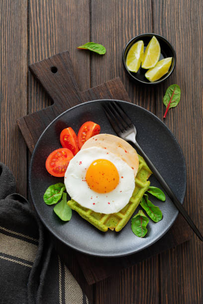 istock Fried egg with cheese, tomatoes, chard, and waffle with spinach on black ceramic plate on brown old wooden background. Sandwich for breakfast. Selective focus. Top view. Copy space. 982488156