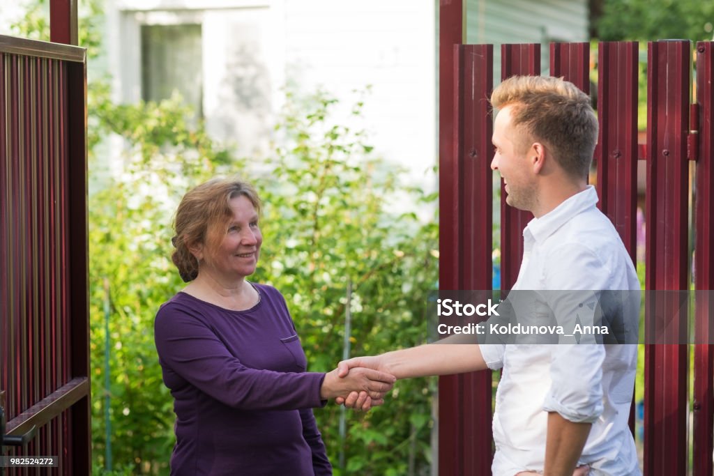 Neighbors discuss the news, standing at the fence. An elderly woman talking with a young man Neighbors discuss the news, standing at the fence. An elderly woman talking with a young man. They are satisfied with this meeting Neighbor Stock Photo