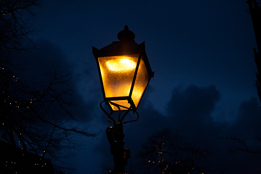 Old lantern illuminates the street with bright yellow glow, dark blue night sky background.