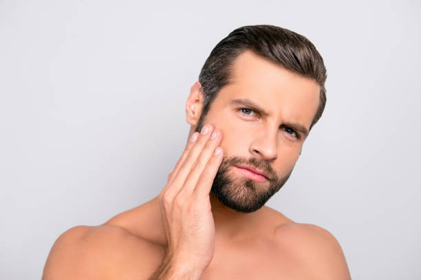 istock Attractive, unhappy, displeased, harsh, virile macho having problem with face, holding hand on cheek, he needs lotion, cream, mask to make soft, smooth, clear, clean skin, isolated on gray background 982669246