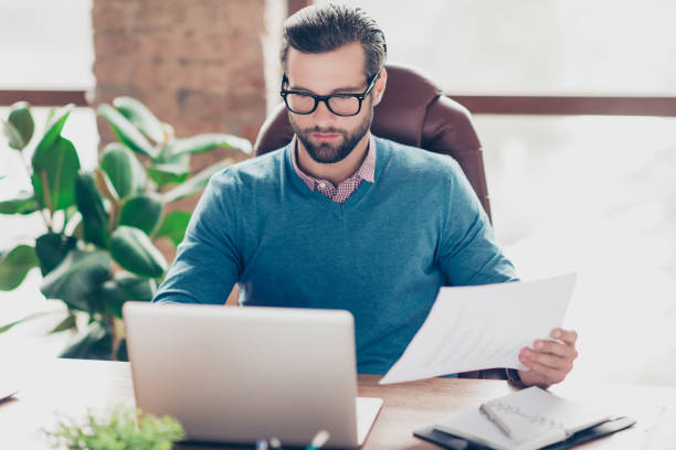 istock Stunning, virile, harsh, manly, professional manager in shirt and pullover holding paper in hand, looking at screen of computer, sitting on armchair at desk in workstation 982702378