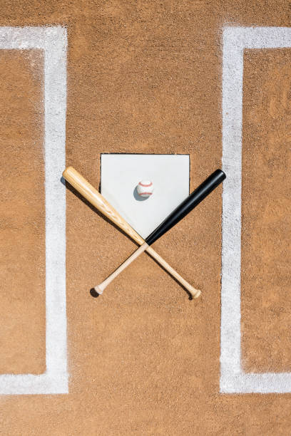 Looking down on a baseball and two wooden bats on home plate Looking down on a baseball and a pair of wooden bats crossing as they sit on home plate with hints of the white lines of the batter's box. home plate stock pictures, royalty-free photos & images