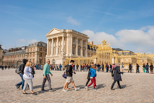 Versailles / France - May 17, 2018: People walk across the plaza at the entrance to the Palace of Versailles, in front of the long line of people waiting to get in. Millions visit Versailles yearly.