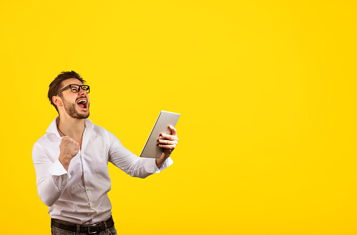 Cheerful bright hipster man holding fist up while screaming with happiness of win on yellow background and holding tablet