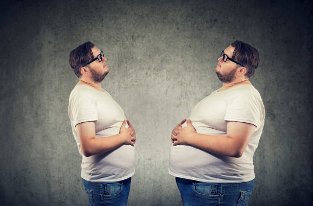 Young chubby man looking at fat himself feeling bloated. Young chubby man looking at fat himself feeling bloated. Diet and nutrition choice  healthy lifestyle concept before and after weight loss stock pictures, royalty-free photos & images