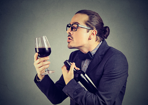 Formal ironic man holding bottle and glass of red wine looking with love to alcohol on gray background