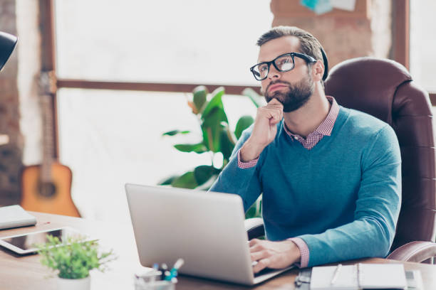 istock Stunning, virile, harsh, professional journalist in shirt and pullover trying to find solution, holding hand on chin with thoughtful expression, sitting on armchair at desk in workplace, using computer for work 982816122