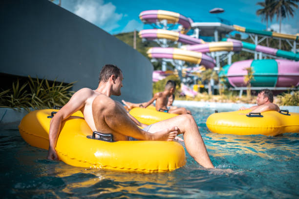 Smiling men relaxing on yellow inflatable ring at water park. They are floating in swimming pool. They are enjoying summer vacation. Water slide is in the back.