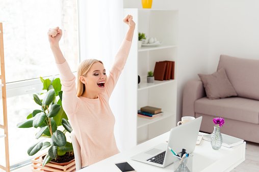 Portrait of successful positive girl celebrating pass exams with raised fists looking at screen of computer having great results sitting in modern white room indoor