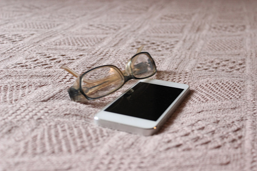 One old eyeglasses with a white mobile phone on the bed in the house