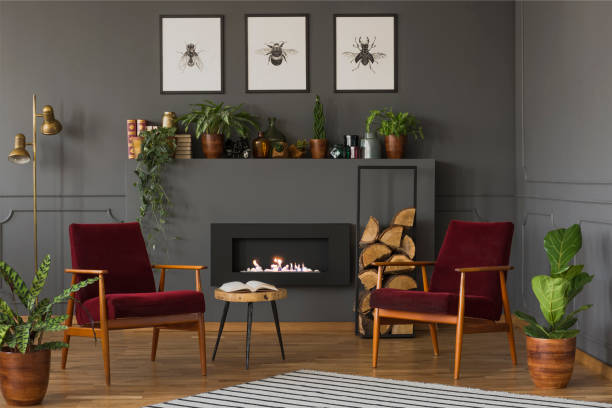 istock Plants next to dark red wooden armchairs in flat interior with fireplace under posters. Real photo 982988192