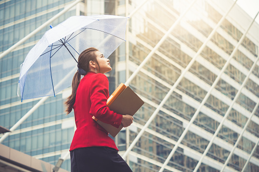 Young beautiful Asian business woman holding umbrella and documents walking to office for working under sunlight and hot weather.
