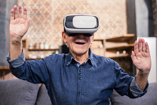 New hobby. Excited smilin elderly man sitting at home in VR glasses and expressing joy