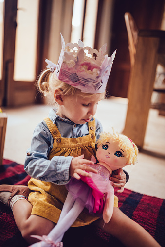 Sad little girl with princess crown holding doll during playtime at nursery daycare