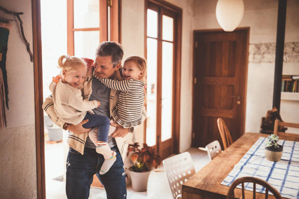 Happy father holding little daughters in his arms at home Loving father holding and embracing little cheerful daughters after returning home from work returning home from work stock pictures, royalty-free photos & images