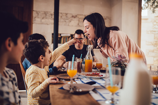 Happy son feeding mother pancake during breakfast with family at home in the morning