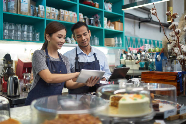 Owner showing digital tablet to colleague in cafe Owner showing digital tablet to colleague. Man and woman are calculating finance. They are wearing aprons in cafeteria. small business malaysia stock pictures, royalty-free photos & images