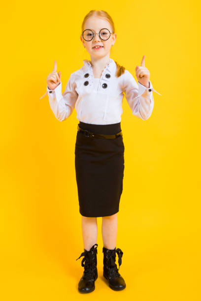 Girl with red hair on a yellow background. A charming girl in transparent glasses points her fingers up. Portrait of a beautiful girl in a white blouse and black skirt. see through leggings stock pictures, royalty-free photos & images