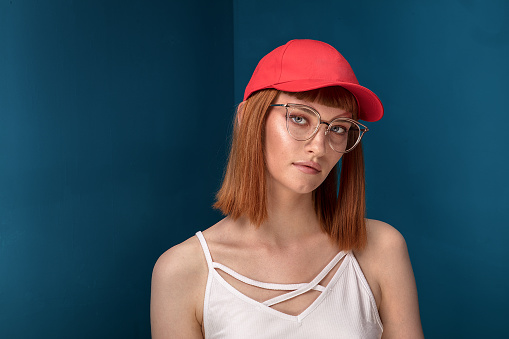 Portrait of attractive young redhead woman with fashionable eyeglasses and cap. Girl looking at camera. Blue background.