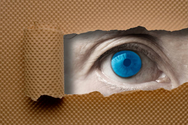 istock Blue eye looking through from a ripped hole in brown paper 983783428