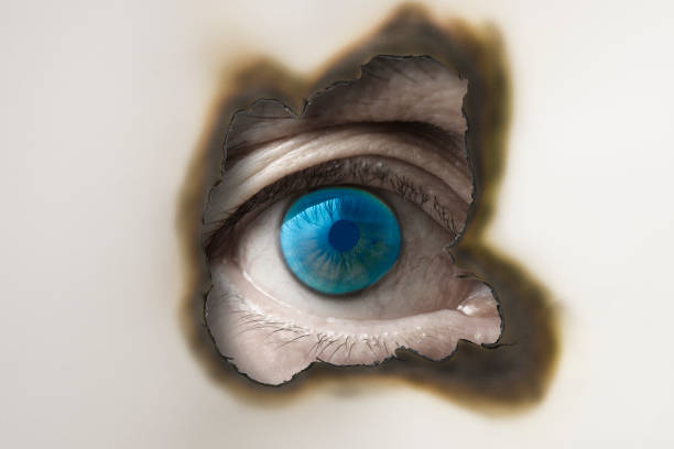istock Blue eye looking through from a burnt hole in white paper 983783436
