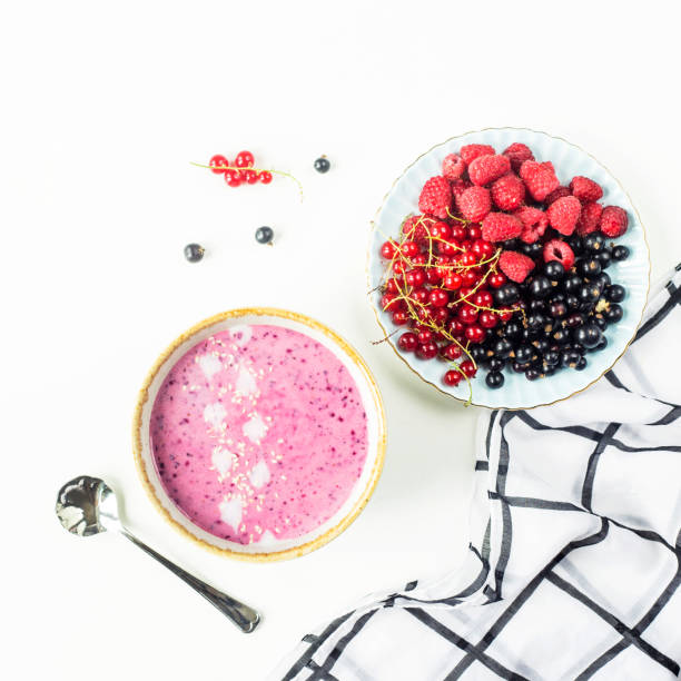 Berry smoothie in a bowl next to a plate with raspberries and black and red currants on a white table with a black and white checkered napkin. Healthy food concept. Top view